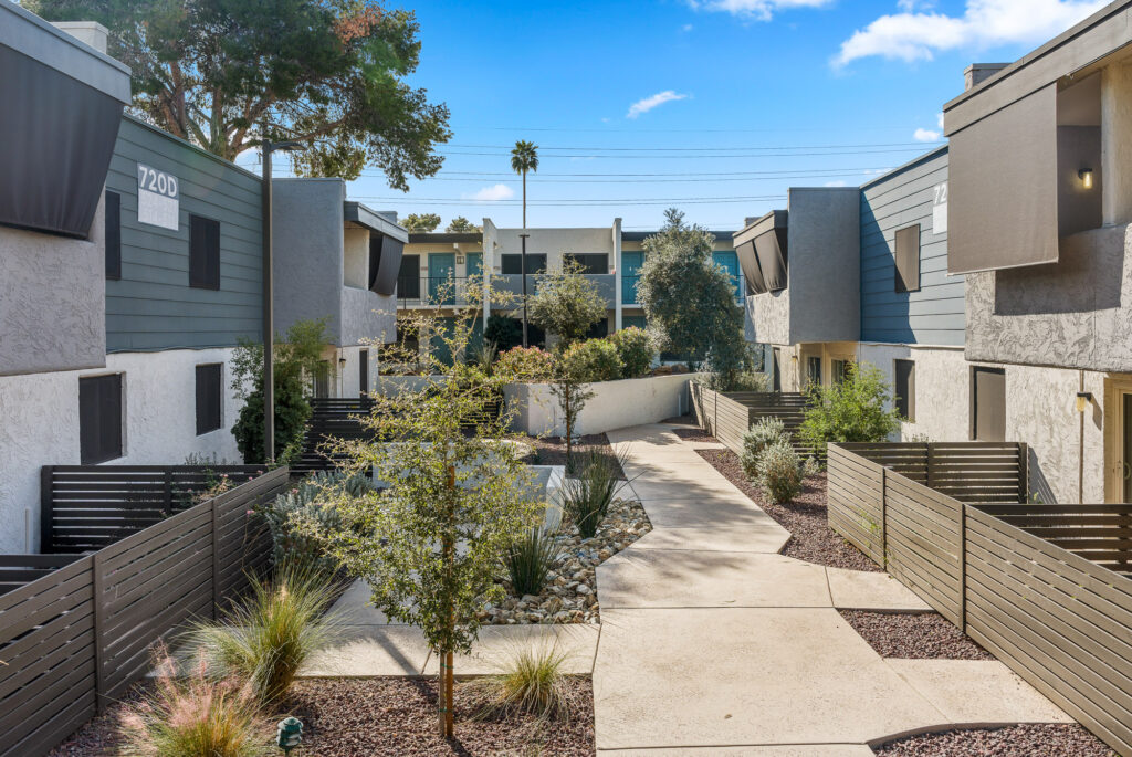 A clean, modern apartment complex features a central walkway lined with plants, trees, and rocks, flanked by two-story buildings with gray and white exteriors, patios, and fences under a bright, sunny sky.