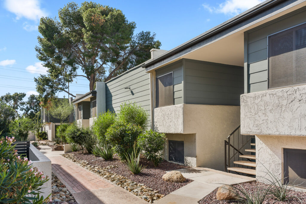 Modern apartment buildings with green siding and stucco exteriors, surrounded by landscaped walkways, shrubs, and trees under a blue sky. Steps lead to the entrances of each unit.