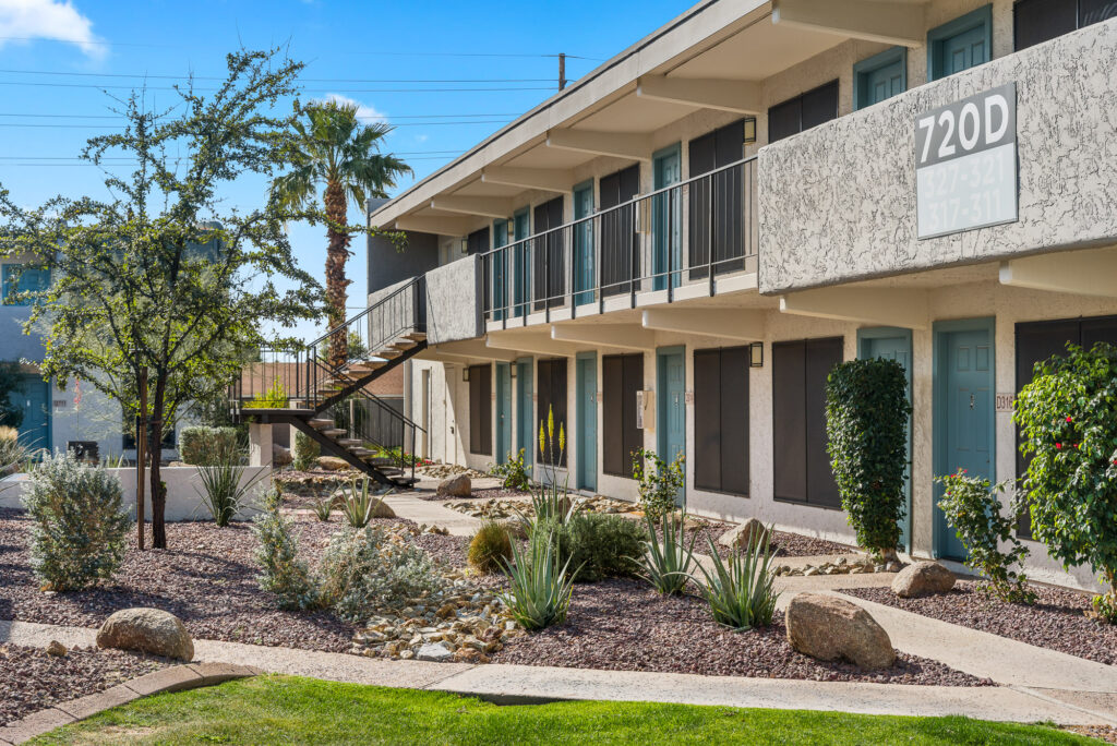 A modern two-story apartment building with blue doors and exterior staircases. The landscaping features desert plants, rocks, and gravel, with a small patch of green grass in the foreground.