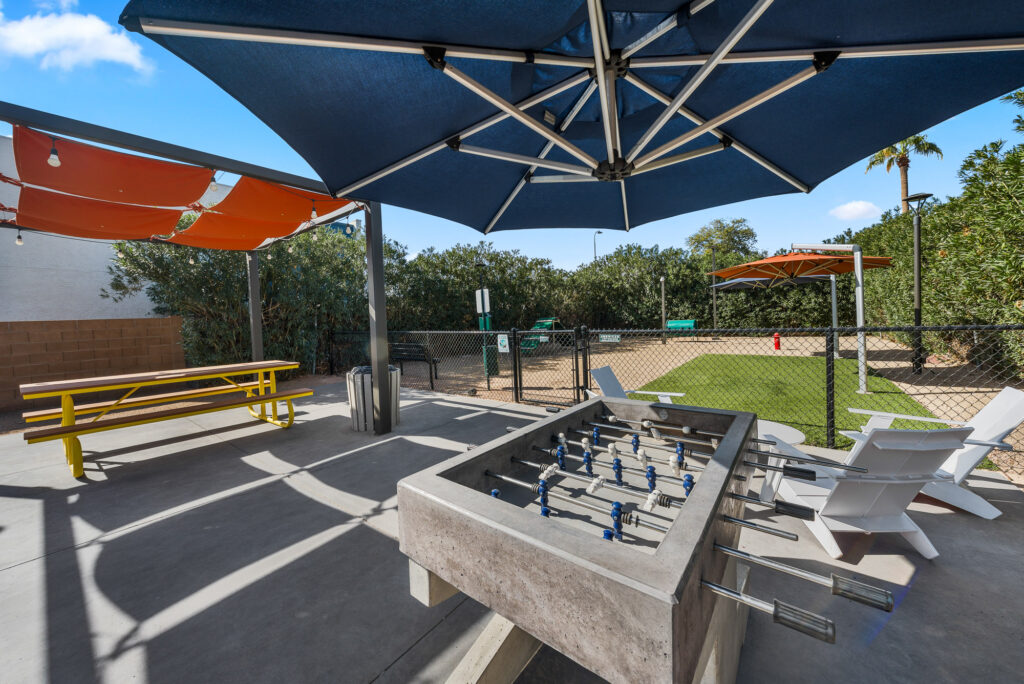 Outdoor recreation area with a foosball table, yellow picnic table, white Adirondack chairs, and large sunshades. A fenced-in sand area and greenery are visible in the background under a blue sky.