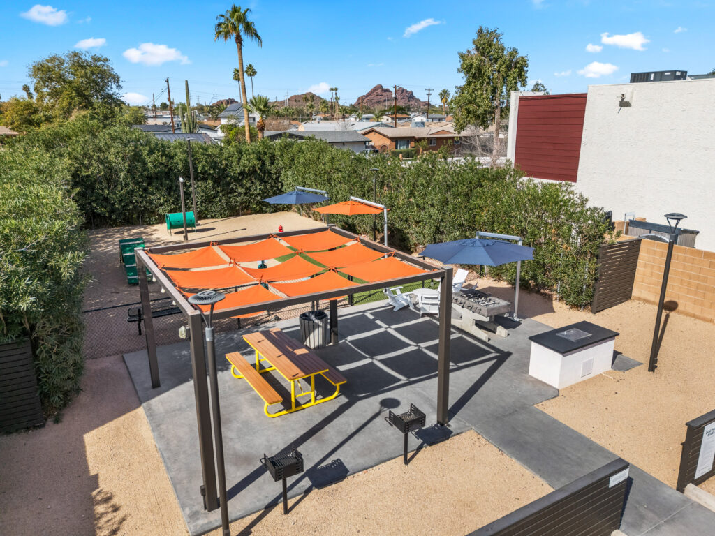 Outdoor patio area with orange shade covers, picnic tables, grills, umbrellas, and a fire pit, surrounded by greenery and a tall privacy fence, with mountains visible in the background under a blue sky.