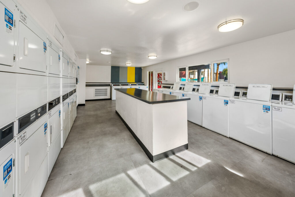 Spacious, clean laundromat with rows of white washing machines and dryers along the walls, a large central folding table, bright overhead lights, and windows letting in natural light.