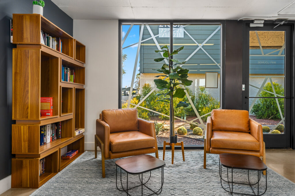 A modern lounge area with two brown leather armchairs, a tall plant, two small wooden tables, a gray rug, a wooden bookshelf with books, and large windows showing an outdoor landscape.