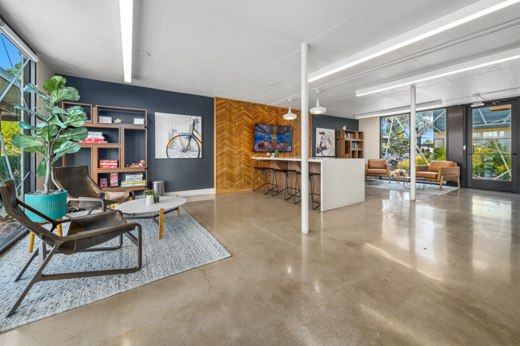 Modern, open-concept lounge with polished concrete floors, stylish seating, a coffee table, wall art, a TV, bar stools at a counter, and large windows letting in natural light. Indoor plants add greenery to the space.