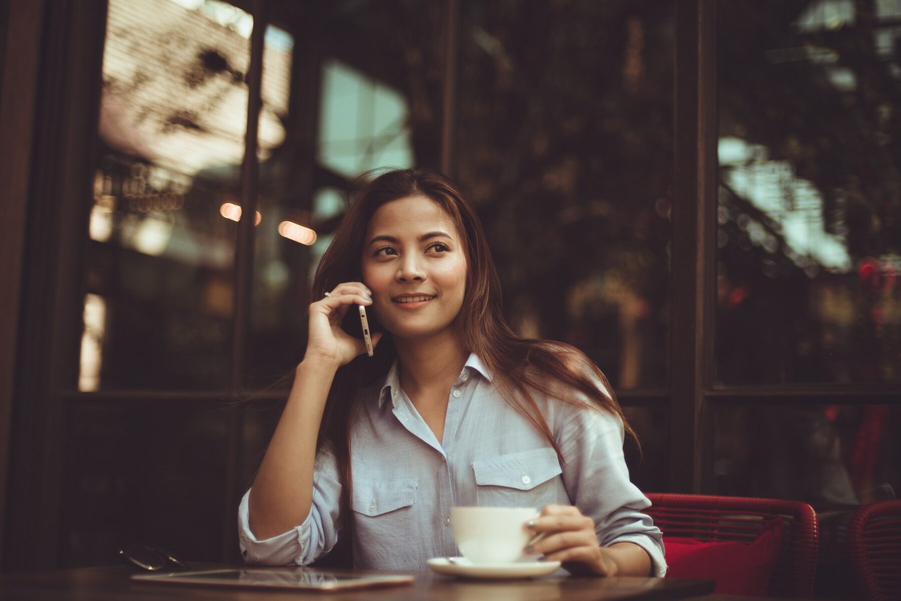 A woman sits at a table in a café, smiling while talking on a smartphone and holding a cup of coffee. She is wearing a light blue shirt, and a tablet is on the table in front of her.